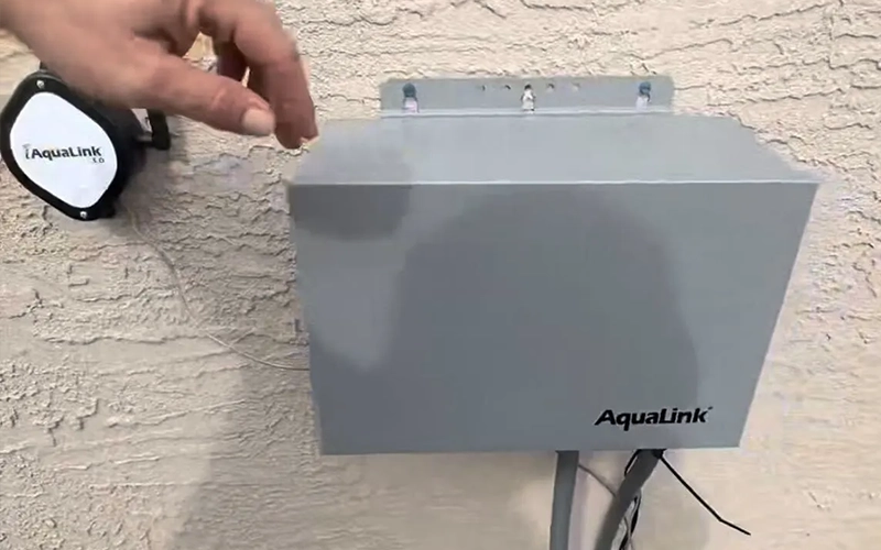 AquaLink pool automation control box mounted on an exterior stucco wall beside an iAquaLink wireless device, with wiring and conduit visible below.
