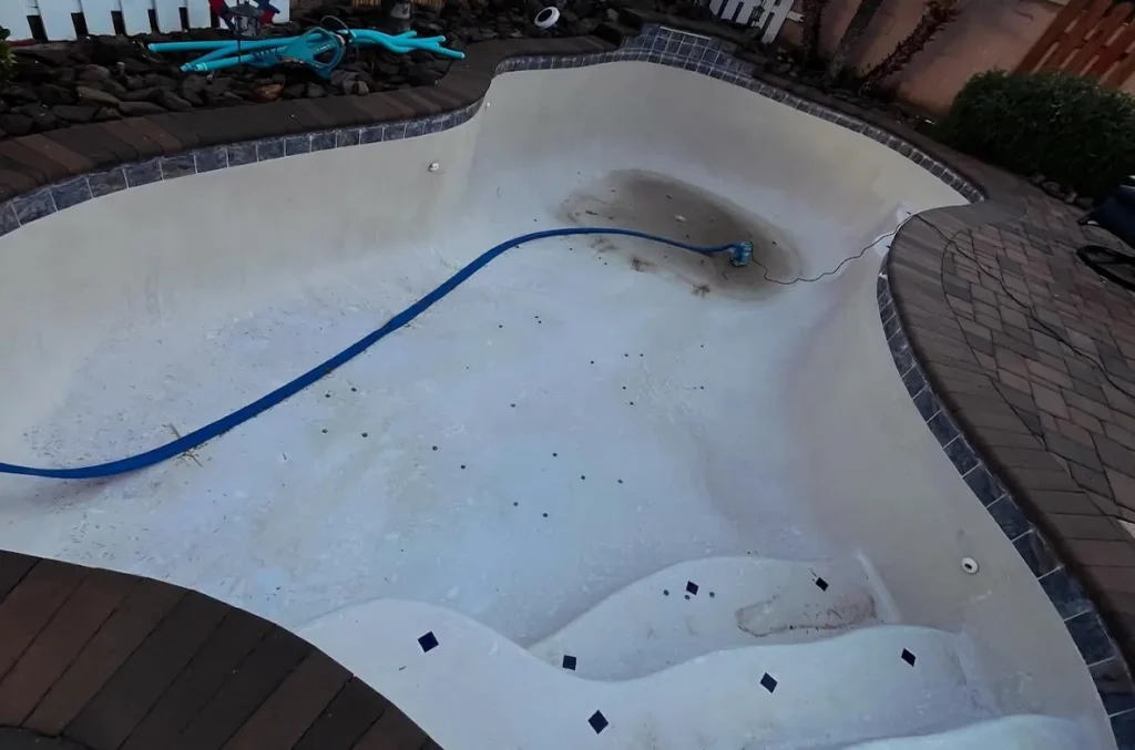 Drained swimming pool with debris and a submersible pump in place before professional pool cleaning and wash service.