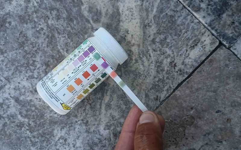 Hand holding a pool test strip next to a water chemistry chart to check chlorine, pH, and alkalinity levels.