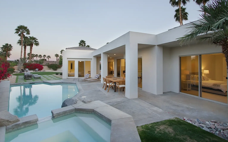 Modern white desert-style home with a backyard swimming pool, spa, patio dining area, and palm trees at dusk.