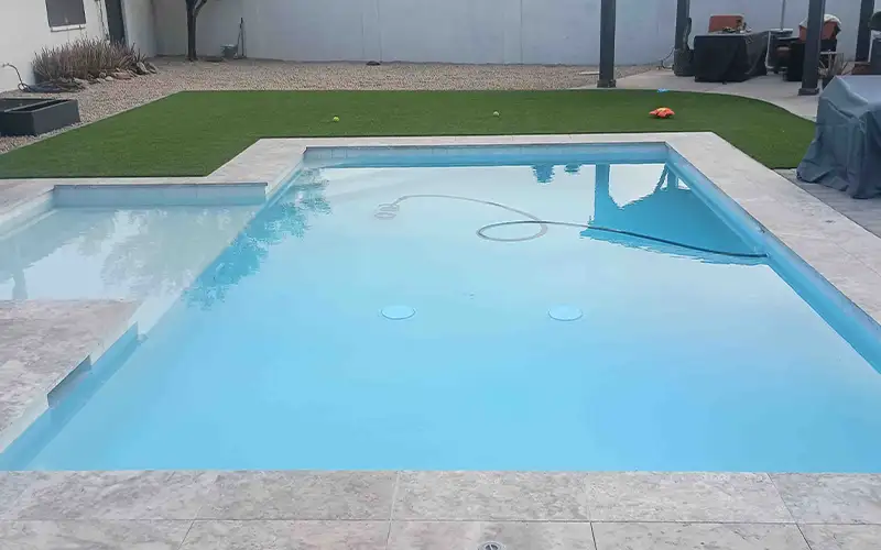 Clean rectangular swimming pool with light blue water, stone patio, artificial grass, and pool vacuum hose
